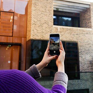 The German language courses building of Goethe-Institut Irland is pictured. The building is made of glass on the left and is orange. On the right it is made of sandstone coloured bricks and has large windows. From behind, a woman photographs the building using her smartphone. Her jumper is purple