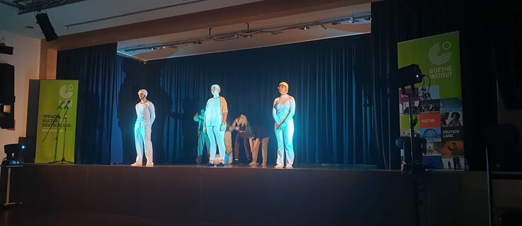 Tres jóvenes vestidos completamente de blanco se sitúan al frente con luces azules que los iluminan. En el fondo, cuatro personas vestidas de beige están en movimiento, claramente trabajando duro en algo que tienen delante. 