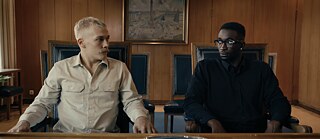 Ein Foto eines Filmausschnitts mit braunem Hintergrund. Zwei junge Männer – der rechte Mann ist Schwarz und trägt ein schwarzes Hemd, der linke ist Weiß und trägt ein beiges Hemd – sitzen nebeneinander in einem schicken Behördensaal mit braunen Holzwänden auf zwei eleganten Stühlen mit schwarzem Lederbezug. Sie tauschen einen leicht irritierten, scheinbar unschuldigen Blick aus. Zwischen ihnen im Hintergrund hängt ein Gemälde der Place de la Bastille in Paris.