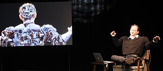 Ein Mann spricht auf der Bühne, neben ihm auf einen Bildschirm ist eine computererzeugte Figur