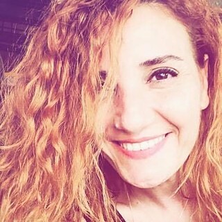 The image shows a close-up portrait of the Cypriot poet, playwright and director Aliye Ummanel. Ummanel smiles at the camera and her red curls cover part of her face. The basic tone and light incidence of the photo is red-yellow.