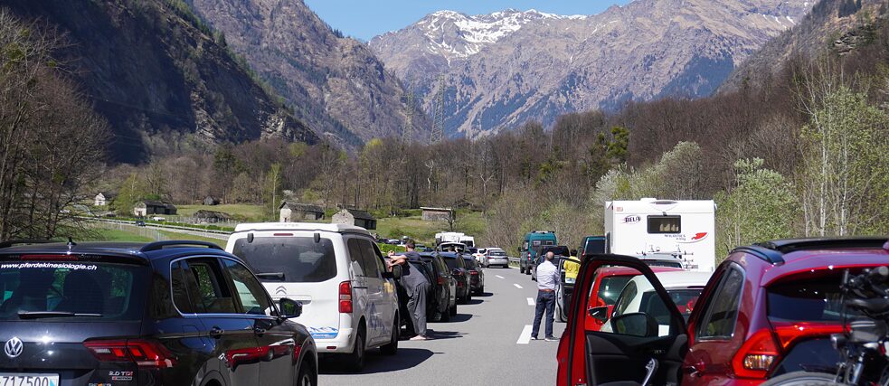 Two queues of vehicles are standing on a highway. Some people have left their cars while waiting. There are mountains in the background.
