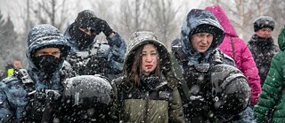Several people, some wearing helmets, apparently policemen lead away a young woman; it is snowing