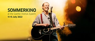 Resimde, Gundermann filminin ana karakteri Gerhard Gundermann, elinde bir gitarla seyircilere gülümseyerek sahnede dururken görülüyor. Arka planda sahne sisi ve sarı ışıklar birbirine karışmış.