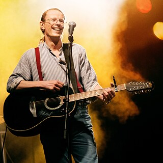 Resimde, Gundermann filminin ana karakteri Gerhard Gundermann, elinde bir gitarla seyircilere gülümseyerek sahnede dururken görülüyor. Arka planda sahne sisi ve sarı ışıklar birbirine karışmış.