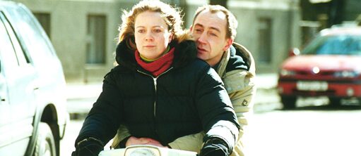 The image of the film shows a couple on a moped. A woman in a black winter jacket, red scarf and blond hair sits in front and steers while looking intently at the road. Behind her, holding on tightly to her, sits a balding man in a beige winter jacket, looking forlornly at the road in front of them.