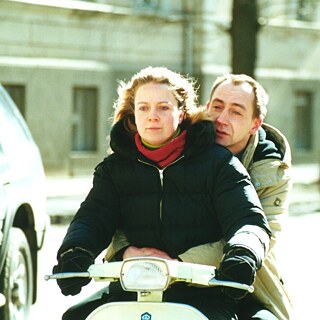 Bir film sahnesinde bir moped üzerinde bir çift görülüyor. Mopedin önünde siyah kışlık ceket giymiş, kırmızı atkılı, sarı saçlı bir kadın oturuyor ve yola konsantre olmuş bir şekilde direksiyonu kullanıyor. Bej rengi kışlık ceket giymiş, alnı açılmış bir adam kadının arkasında oturuyor, kadına sıkıca sarılmış ve boş gözlerle önlerindeki yola bakıyor.