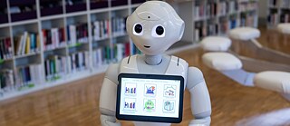 A robot stands in a library; on its screen, different possibilities for use are displayed.