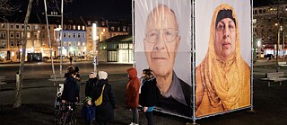 Several people attend an evening public exhibition of large portrait photos in a public area