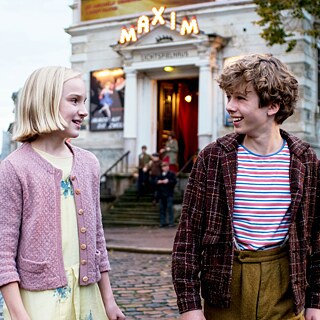 Στη φωτογραφία διακρίνονται δύο παιδιά σε ημι-κοντινό πλάνο να κοιτάζονται και να συνομιλούν σε μια πολυσύχναστη πλατεία. Το αγόρι, στα δεξιά της εικόνας με τις καφέ κοντές μπούκλες, γελάει ζωηρά. Η κοπέλα, ανοιχτόξανθη με μαλλιά μέχρι το πηγούνι, τον κοιτάζει με ενδιαφέρον και φιλικότητα.