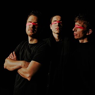 Three men in black stand next to each other. Their clothes blend with the pitch-black background. Their eyes are covered with a pink tape.