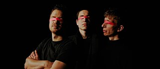Three men in black stand next to each other. Their clothes blend with the pitch-black background. Their eyes are covered with a pink tape.