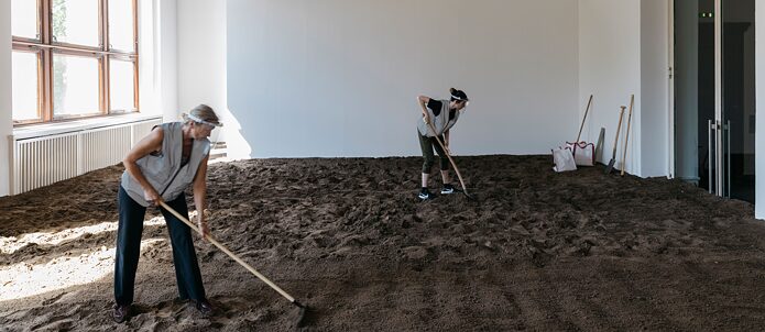 2 people rake the soil on the floor of a gallery space