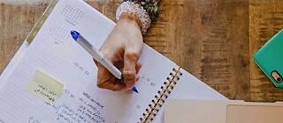 Eine Frau schreibt in ein Heft. Auf dem Tisch befinden sich ein Laptop, ein Handy, ein Hut und ein Trinkbecher mit Limonade.