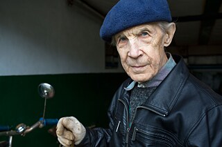Ein älterer Mann mit Lederjacke und blauer Schiebermütze steht in einer Garage. Im Hintergrund steht ein altes Fahrrad.