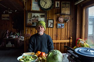 Ein Mann mit grüner Mütze sitzt an einem Tisch, auf dem Lebensmittel liegen. An der Holzwand im Hintergrund hängen Familienbilder.