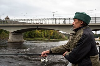 Ein Mann steht an einem Fluss und hält eine Angel in der Hand. Er trägt eine grüne Mütze und hat eine Zigarette im Mund.