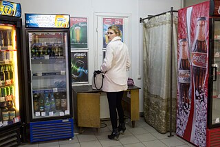 Eine junge Frau mit rosa Mantel steht in einem Laden an einem Tisch und schaut über ihre Schulter. Neben ihr befinden sich Getränkekühlschränke und eine Umkleidekabine.