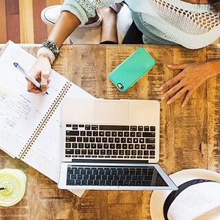 Eine Frau schreibt in ein Heft. Auf dem Tisch befinden sich ein Laptop, ein Handy, ein Hut und ein Trinkbecher mit Limonade.