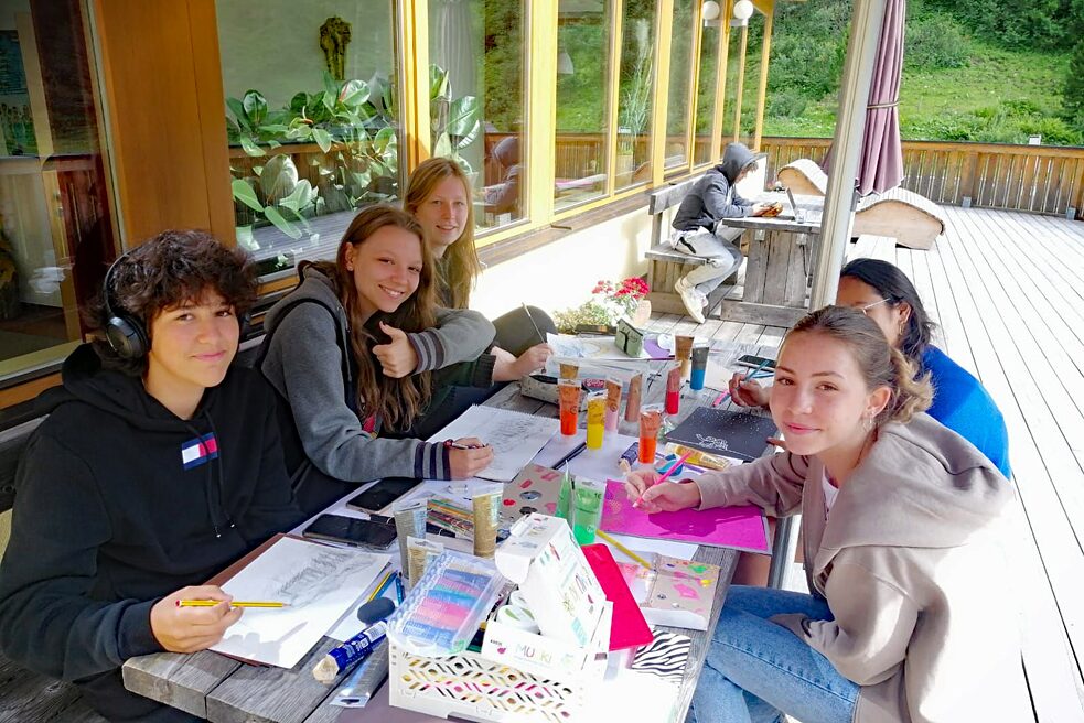 Jugendliche vor einer Almhütte, Bastel- und Malmaterial liegen auf dem Tisch.