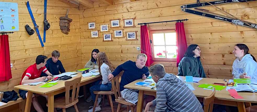 Jugendliche in einer Almhütte, vor ihnen lieben Lernmaterialien auf den Tischen.