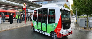 A small, autonomously driving electric bus at the city railway station. There are a few people waiting at the bus stop.
