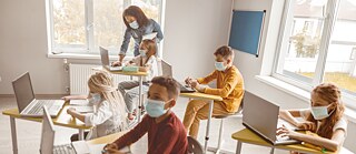 Maske tragende Schülerinnen und Schüler sitzen an Einzeltischen im Klassenraum. Alle haben einen Laptop.