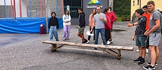 Jugendliche und ihr Lehrer stehen auf einem Hof vor einer Holzbank.