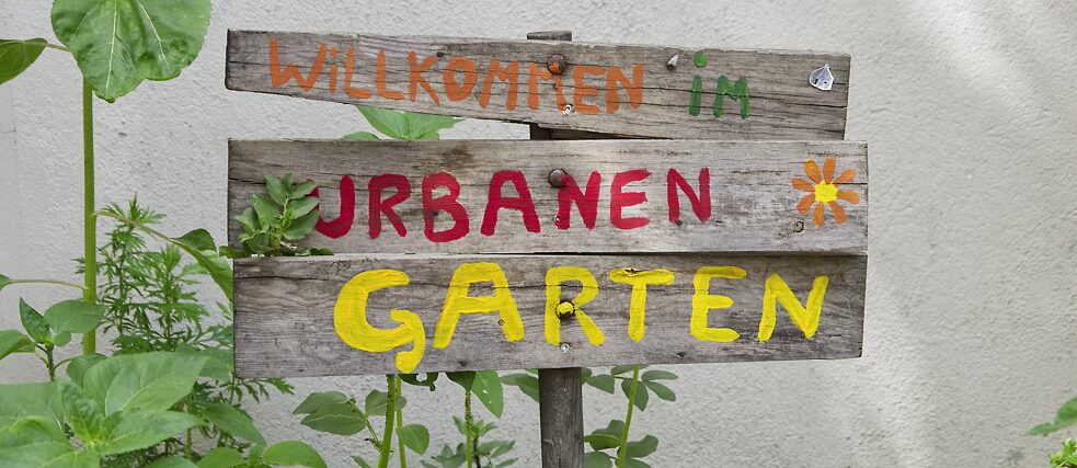 „Willkommen im urbanen Garten“, Begrüßungstext auf einem Holzschild.