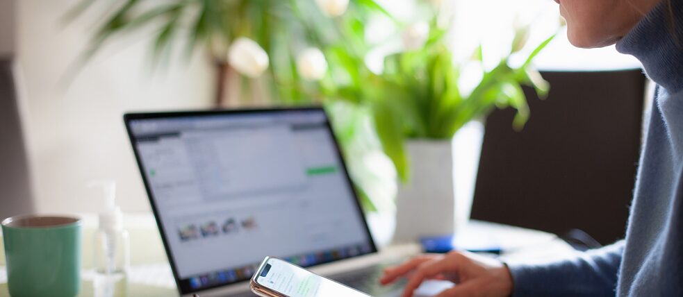 Eine Geschäftsfrau sitzt zu Hause am Laptop und überprüft gerade die Textnachrichten auf ihrem Smartphone. Vor ihr steht eine Kaffeetasse.