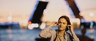 Ein junger Mann mit Kopfhörern sitzt am Flussufer vor einer geöffneten Brücke in St. Petersburg