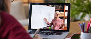 Learner at a video conference. She takes notes. The instructor can be seen on the screen. She is standing in front of a whiteboard. 