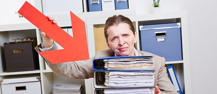 A woman sitting in an office pointing with a red arrow at a stack of files and grimacing.