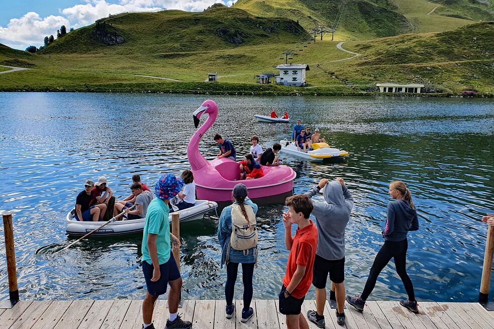 Jugendliche auf einem Steg, im Hintergrund ein Bergsee und Tretboote.