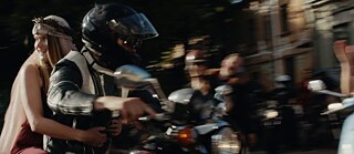 Zu sehen sind zwei Personen auf einem Motorrad. Die vordere Person trägt einen schwarzen Motorradhelm, die  hintere Person trägt keinen Helm und schaut lächelnd in die Richtung, aus der das Motorrad kommt.