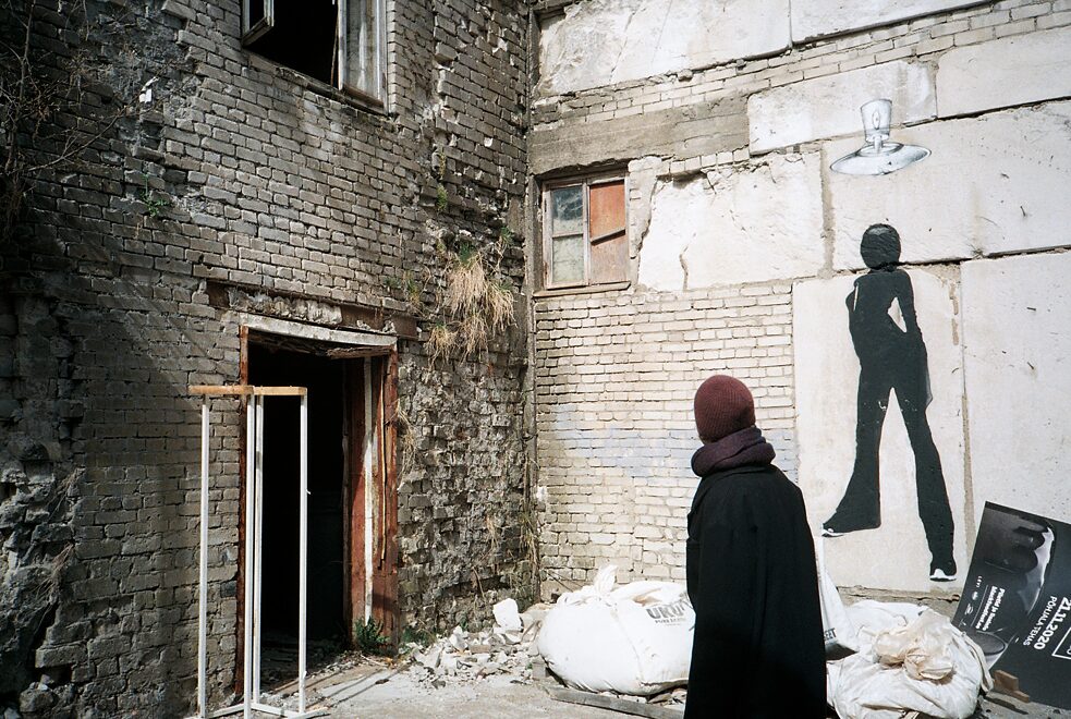 Eine Person in schwarzem Mantel steht vor einem alten Gebäude, auf dem die Silhouette einer Frau aufgemalt ist.