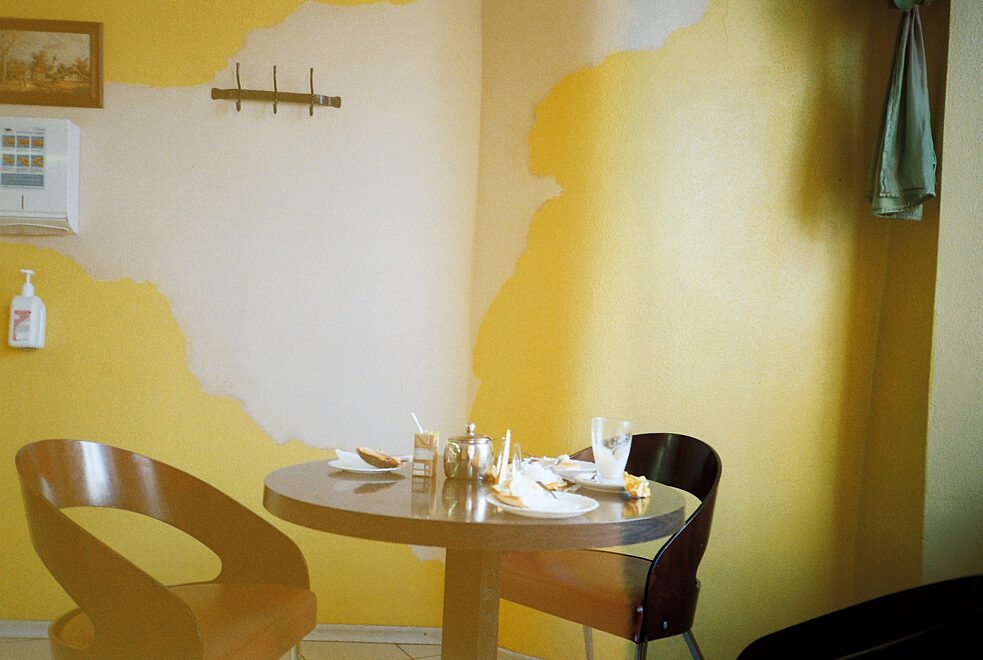 Ein verlassener Tisch in einem Café, auf dem noch leeres Geschirr und Müll ist. Die Wand im Hintergrund ist gelb.