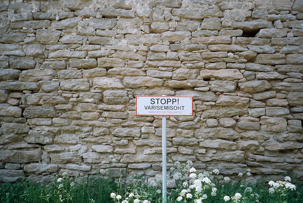 Ein Schild mit der Aufschrift „Stopp! Varisemisoht. Danger of Collapse.“ steht vor einer Mauer.