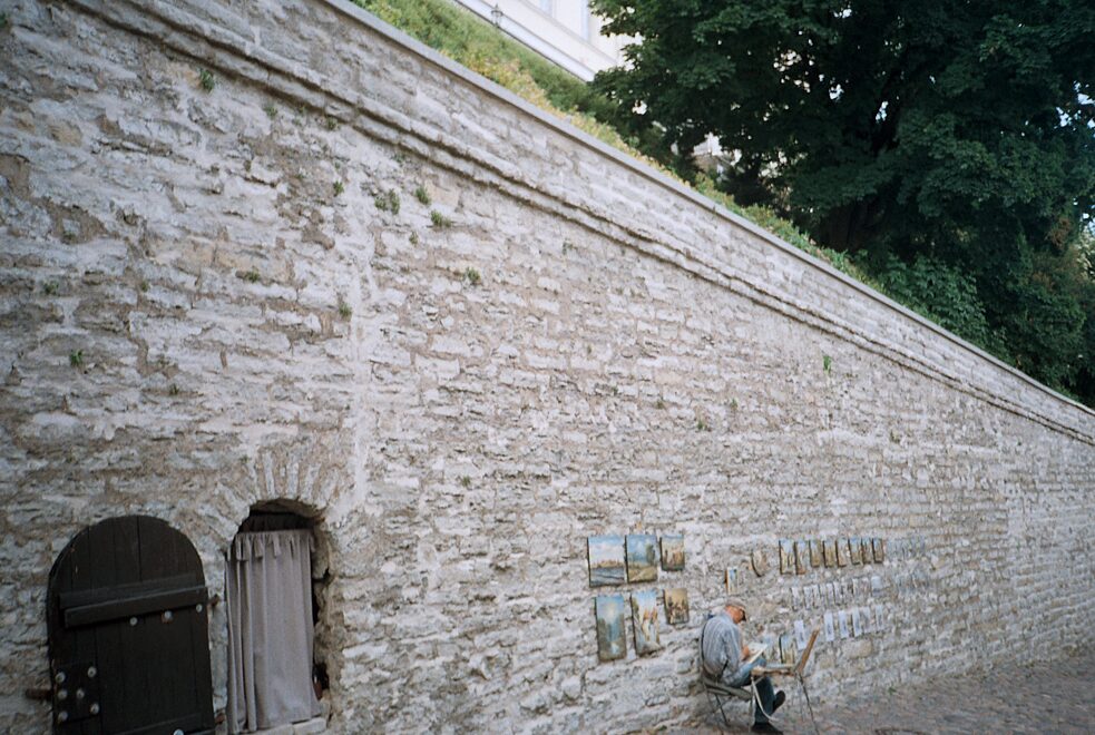 Мужчина сидит перед высокой городской стеной и рисует, его картины выставлены на стене.