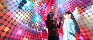Two women in front of a colourful background one with VR glasses 