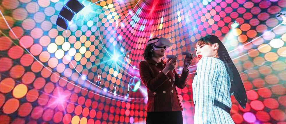 Zwei Frauen vor einem bunten Hintergrund eine mit VR- Brille 