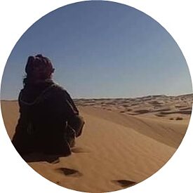 Une femme est assise sur une dune de sable, dos à la caméra.
