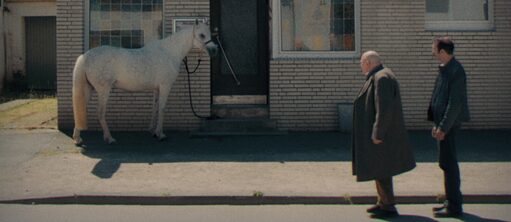 Die beiden Komissare hintereinander stehend im Profil rechts vorne im Bild vor der Hausfassade einer Bar, links im Bild ein weißes Pferd, angeleint an der Tür der Bar. 