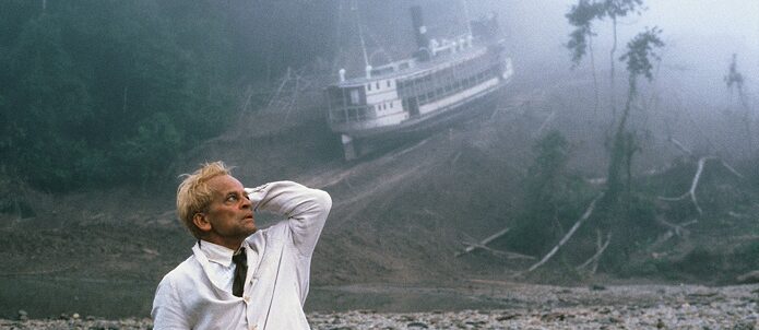 Szene aus dem Film „Fitzcarrald“. Ein Mann in einem weißen Anzug mit einem Hut in der Hand hält den Arm hinter den Kopf und schaut seitlich nach oben. Im Hintergrund liegt ein Dampfschiff auf der Hälfte eines Berges.