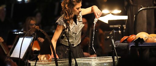 Suyla dolu iki büyük cam çanağın arkasında uzun saçlı kumral genç bir kadın duruyor. Küçük bir cam şişedeki suyu çanaklardan birine boşaltıyor. Arkasında flu görünen bir yaylı çalgılar orkestrası bulunuyor.