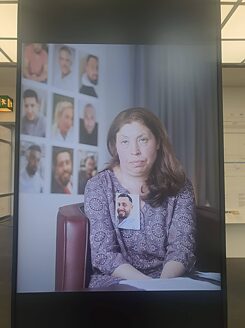 A vertical screen shows a seated woman with a serious and devastated look, who has a photograph of a man pinned on her blouse. On the wall behind her there are portraits of more people.  