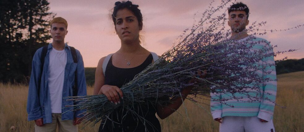 Eine junge Frau mit einem Lavendelstrauß steht in einer ländlichen Landschaft.  An beiden Seiten, etwas weiter hinten stehen zwei  junge Männer.