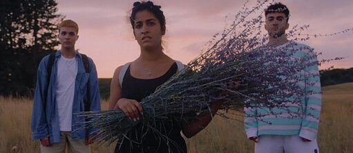 Eine junge Frau mit einem Lavendelstrauß steht in einer ländlichen Landschaft.  An beiden Seiten, etwas weiter hinten stehen zwei  junge Männer.