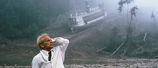 Escena de la película "Fitzcarrald". Un hombre con un traje blanco y un sombrero en la mano sostiene el brazo detrás de la cabeza y mira hacia atrás. En el fondo, un barco de vapor se encuentra a mitad de camino de una montaña.