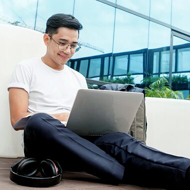 Junger Mann sitzt mit einem Laptop vor einem Firmengebäude